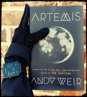 Hand wearing sports gloves and a GPS watch holding a copy of "Artemis".