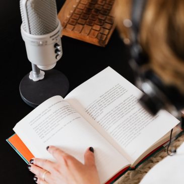 A woman, seen from behind, wearing headphones as she reads a hardcover book into a microphone in front of her.