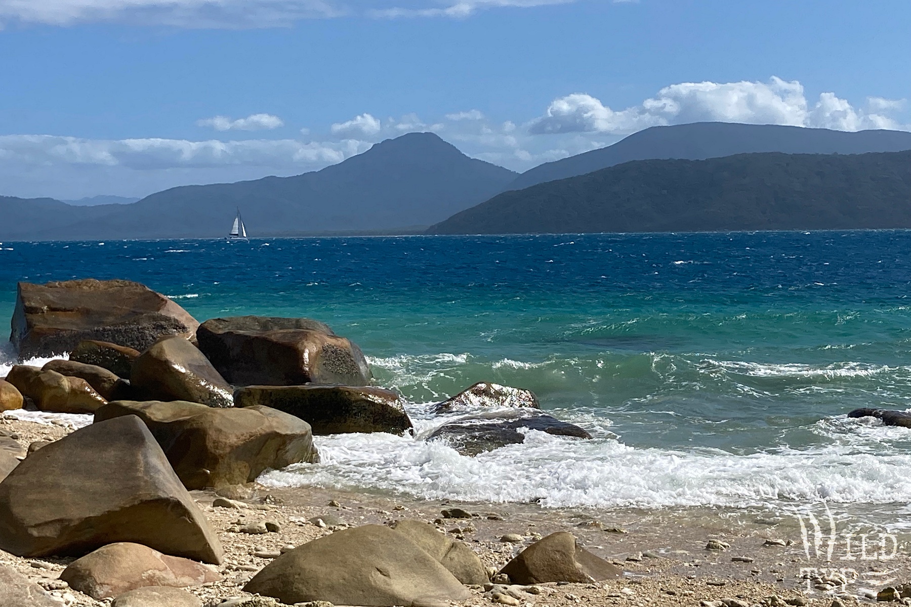 Teal waves lap a rocky beach. Beyond the breakers, the ocean turns deep blue. A far-off sailboat cruises in front of distand jungle hills.