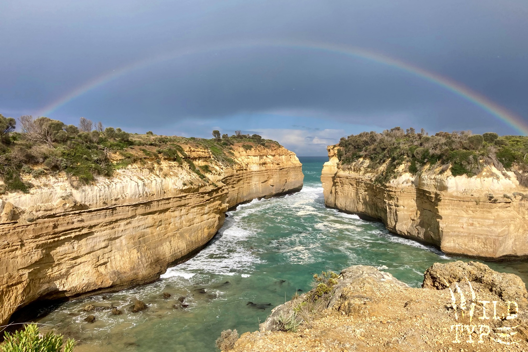A rainbow spans a rugged coastal gorge. Jade-green water slithers between the sandstone cliffs, as if pouring through the rainbow's gate.
