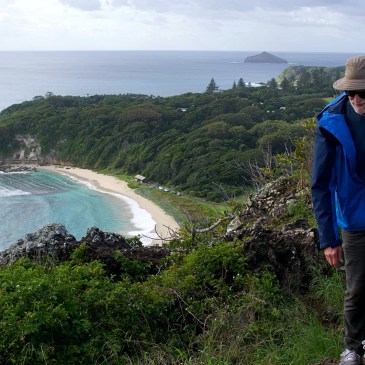 An older man in a blue windbreaker, floppy hat, and sunglasses hikes along a rugged ridge towards the viewer. The edge of an island sweeps away In the distance below: azure water licking a crescent-shaped beach, and jungle giving way to an ocean horizon.