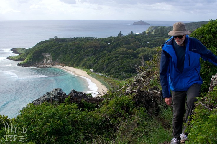 An older man in a blue windbreaker, floppy hat, and sunglasses hikes along a rugged ridge towards the viewer. The edge of an island sweeps away In the distance below: azure water licking a crescent-shaped beach, and jungle giving way to an ocean horizon.