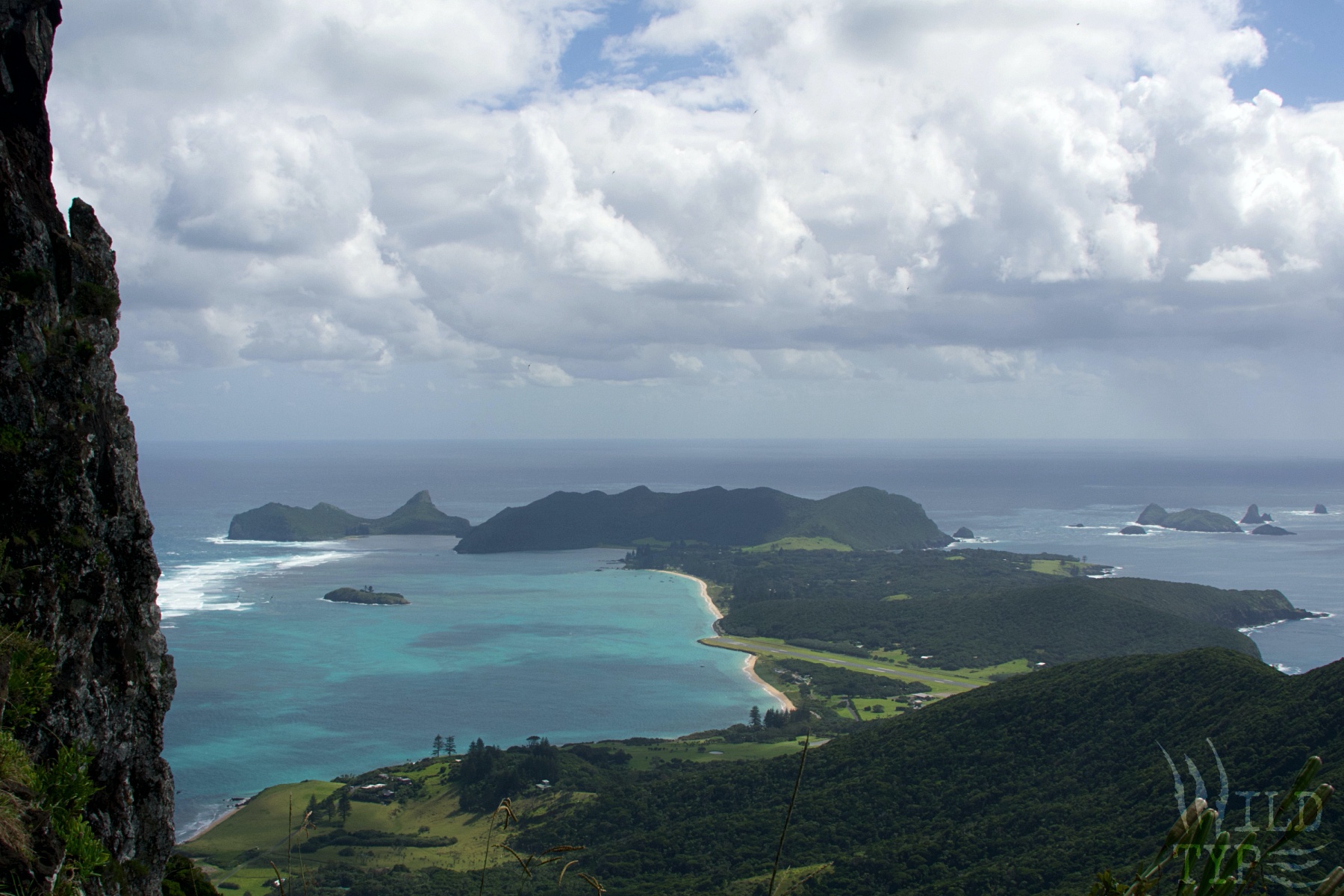 A view from a mountain cave, gazing over a narrow forested island surrounded by bright blue water. Fleecy clouds hang above the island's rugged peaks.