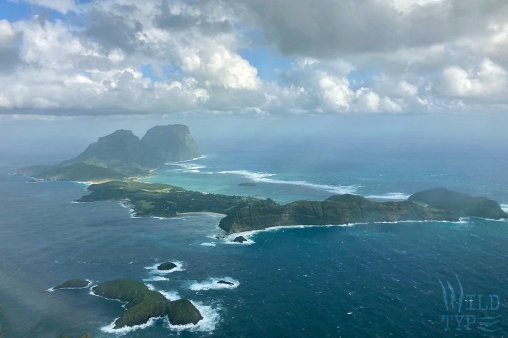 A mountainous green island, ringed with white surf, rises from a turquoise sea.