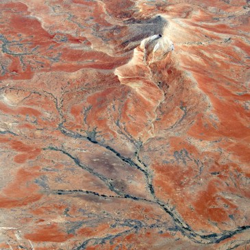 An aerial view of the South Australian outback. Black alluvial veins slither across red earth. Ridges and peaks of white rise up, giving the landscape a crumpled look.